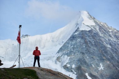 Kırmızı ceketli turist İsviçre bayrağıyla bayrak direğinin yanındaki tepede duruyor, Ober Gabelhorn arka planda, bir tarafı karla kaplı ve diğer tarafı kar değil.