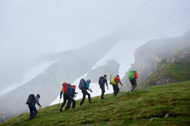 Sırt çantalı erkek yürüyüşçülerin çimenli tepeye tırmanması. Arka planda sisli uçurumla dağlarda yokuş yukarı yürüyen genç turistler. Yürüyüş, seyahat ve sırt çantası kavramı.