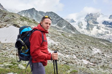 Yürüyüşçü yürüyüşünün yan görüntüsü, yürüyüş çubukları, kameraya bakmak, arka planda karlı güzel dağlar. Dağcılık, dağcılık, Alplerde spor turizmi.