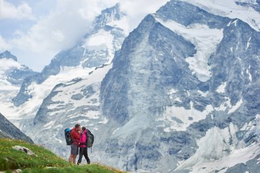 Sırt çantalı sevimli çift öpüşüyor, arka planda güzel dağ manzarası var. Seyahat, turistler birlikte zirveye ulaşıyor. İnanılmaz manzaralı vahşi doğa. Alplerde spor turizmi.