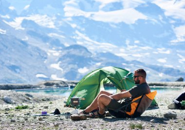 Erkek turist, göl kenarındaki boş kayalık dağlarda çadırın yanında oturuyor ve güneşli yaz gününde akıllı telefon kullanıyor. Doğa kavramı, aletler, yürüyüş ve kamp.