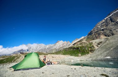 Erkek gezgin arka planında görkemli kayalık tepelerle turist çadırının yanında oturuyor. Akıllı telefon kullanan ve mavi gökyüzünün altındaki dağlarda dinlenen genç adam. Seyahat, yürüyüş ve kamp kavramı.