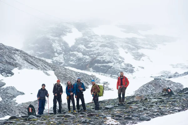バックパックやトレッキングスティックを背景に雪の山と岩の丘の上に立っている男性ハイカーのグループは カメラを見て笑顔 ハイキング 登山の概念 — ストック写真