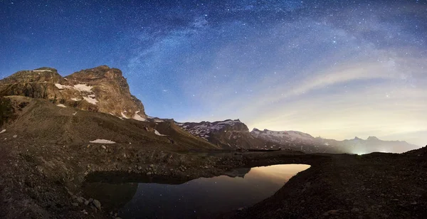 Panoramic view scenery of mountain peak Matterhorn at night under Milky way with shining stars in dark sky. Beautiful mountains area in Alps with high rocky peaks and lake with clear water.