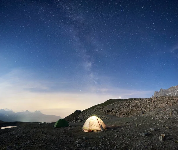 Fantastic view of night camping in mountainous region. Magnificent scenery of tourist tents on rocky hill under beautiful night sky with stars and milky way. Concept of travelling, hiking in Alps.