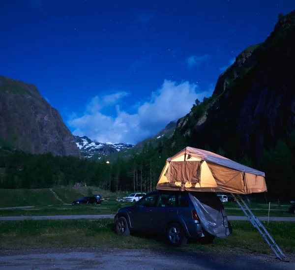 Camping night scene with silver car, majestic hills on background. Beautiful view of blue evening sky over high mountains and SUV with rooftop tent. Concept of travelling, camping and mountain hiking.