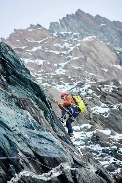 Side view of man climber with backpack using fixed rope to climb high rocky mountain, ascending alpine ridge and trying to reach mountaintop. Concept of mountaineering and alpine rock climbing.