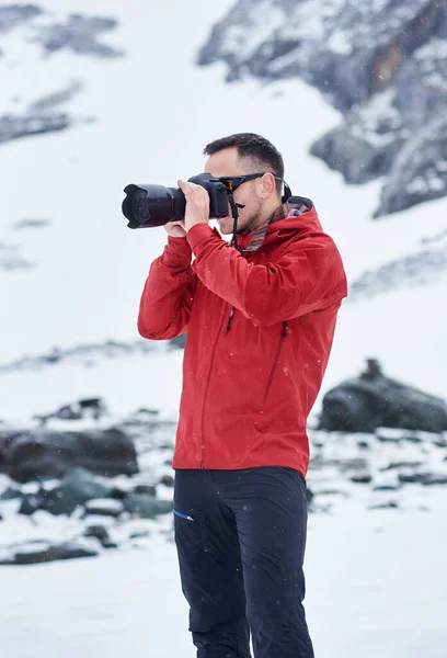Side view of brunet gentleman taking photo with professional camera. Guy wearing red jacket while photographing winter nature. Concept of travelling and professional photography.
