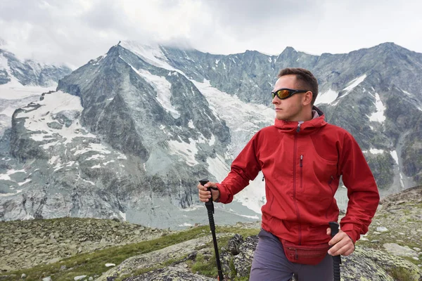 Portrait of a man wearing a red jacket and a backpack, having a walk with hiking poles in the mountains situated in Swiss Alps, rocky ridge is on background