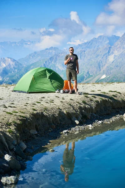 Front view of traveler holding cup of tea while standing by tourist tent, blue lake in mountain valley. Male hiker in shorts reflecting in crystal water. Concept of tourism, hiking and camping.