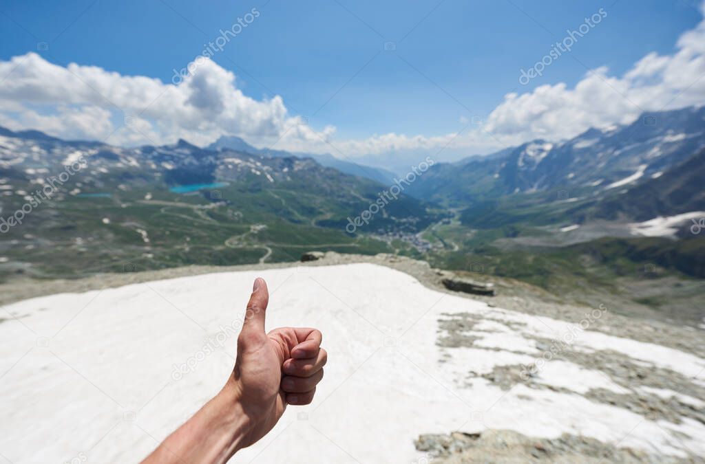 Close up of man hand doing approval gesture with grassy hills and beautiful sky on background. Mountaineer showing thumbs up sign while hiking in mountains. Concept of travelling and mountaineering.