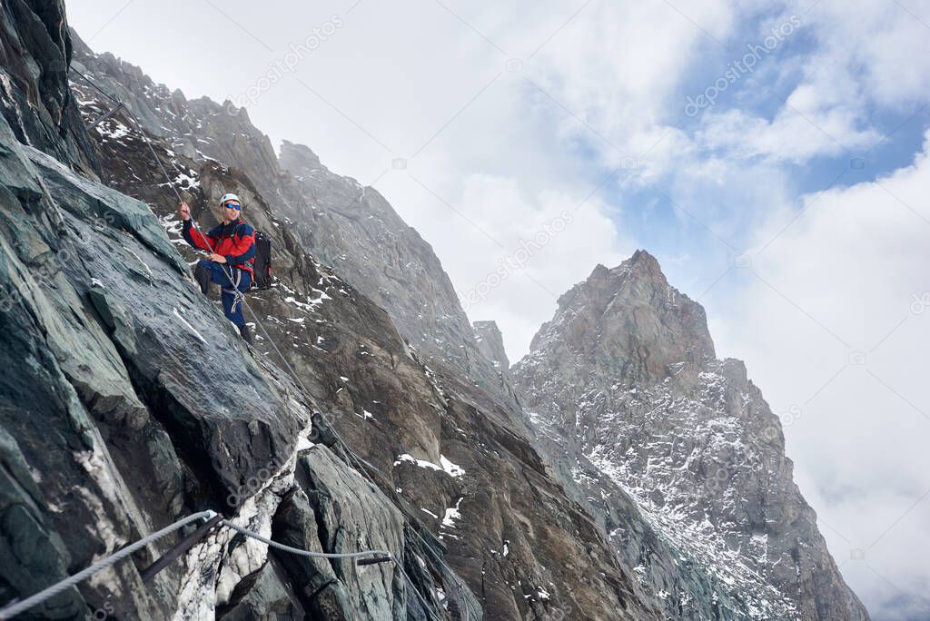 Male alpinist in sunglasses and safety helmet holding fixed rope while climbing mountain. Mountaineer looking aside and smiling while ascending natural rock formation. Concept of alpine rock climbing.