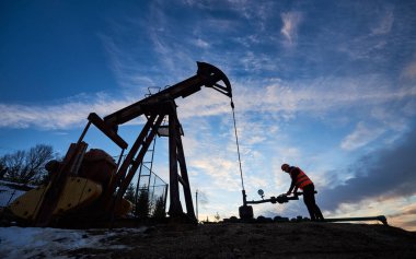Yağ pompalama ünitesini petrol kuyusundan ham petrol çıkarmak için kullanan iş yeleği ve kask giyen petrol operatörünün silüeti.
