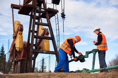 İki petrol işçisi tulum, turuncu yelek ve kask giyiyor, yağ pompası krikosunun yanındaki petrol sahasında çalışıyor, biri boruyu büküyor, diğeri de notlar alıyor.