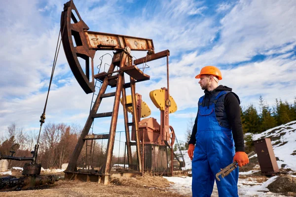 在一个阳光明媚的日子里 身穿蓝色制服 头戴橙色头盔的石油工程师站在一个油田 旁边印着一个平面图 站在抽油机的千斤顶旁边看着钻机 — 图库照片