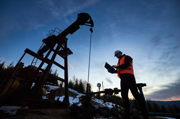 Low angle of petroleum engineer in work vest looking at oil pump rocker-machine and writing on clipboard