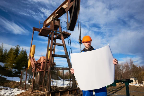 Portrait of an oil worker in a blue uniform and orange helmet standing in an oilfield with a printed plan on a beautiful sunny day, oil pump jack and blue sky on background