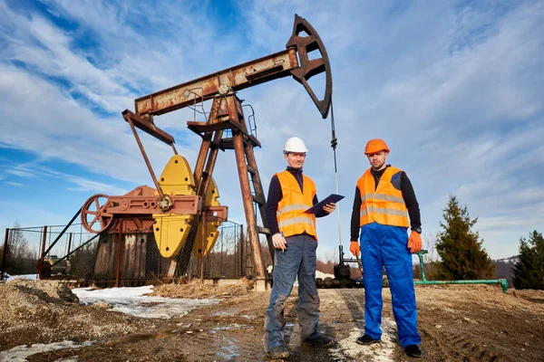 两名头戴钢盔 身穿工作服的石油工人站在油田的土地上 背景是油泵摇臂机和天空 石油工程师持有剪贴板 与油田同事一起工作 — 图库照片
