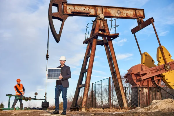 Portrait of smiling businessman in suit jacket, white helmet, holding mini solar panel, showing thumb up. Oil operator working with pump jack on oil field on background. Concept of alternative energy