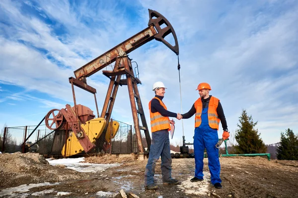 两名男子身穿工作服 身穿橙色背心 头戴钢盔 站在油田旁边 与加油站的千斤顶握手 背景是蓝天 石油工业和石油开采的概念 — 图库照片