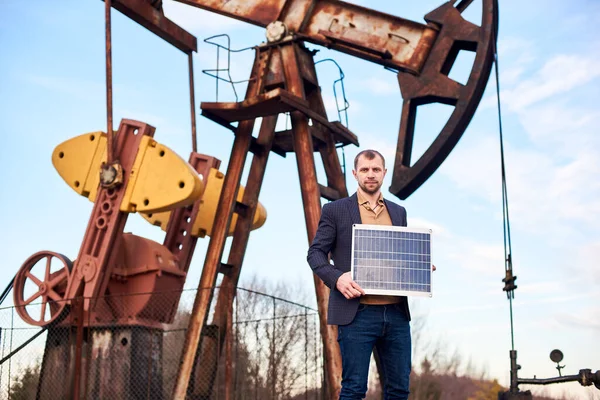 Horizontal portrait of a businessman wearing a sport coat, jeans holding mini solar battery, oil pump jack behind him, concept of alternative source of energy
