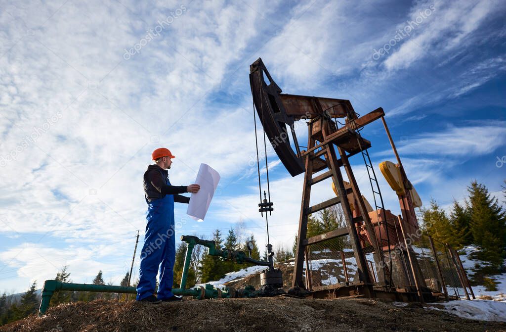 Oil engineer in a blue uniform and orange helmet standing in an oilfield with a printed plan next to the oil pump jack looking at the rig, on a beautiful sunny day