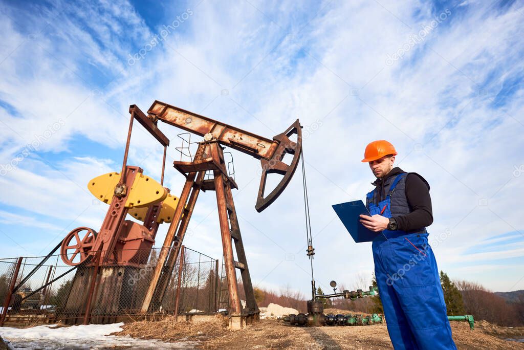 Petroleum engineer in work overalls and helmet holding clipboard, checking oil pumping unit, making notes. Oil worker standing near oil pump jack under beautiful sky. Concept of oil extraction