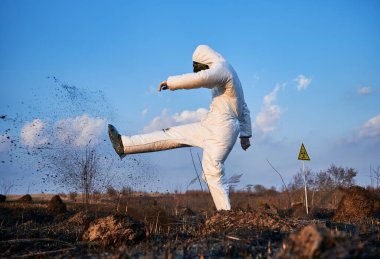 Hayal kırıklığına uğramış bir bilim adamının yatay yan görüntüsü. Beyaz koruyucu bir elbise, gaz maskesi ve eldiven giyiyor. Mavi gökyüzüne karşı yanan arazide toprağı tekmeliyor.