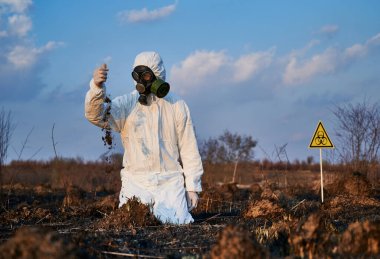 Beyaz koruyucu giysi, gaz maskesi ve eldiven giyen erkek bilim adamı dizlerinin üzerinde karınca yuvalarının arasında duruyor ve elinden toprak döküyor, arkasında biyolojik tehlike işareti var. Ekoloji kavramı, yanmış toprak.