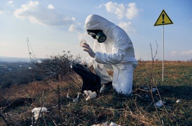 Beyaz koruyucu elbise giyen, gaz maskeli, çömelmiş, güneşli bir günde siyah çöp torbalarına çöp toplayan çevrecilerin yan görüntüsü. Arkasındaki uyarı işareti. Ekoloji kavramı, kirlilik