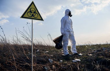 Radyasyon giysisi ve gaz maskeli bir erkek ekolojist çöp torbası tutuyor, çöplerle dolu terk edilmiş bir bölgede biyolojik tehlike uyarısının yanında duruyor. Ekoloji kavramı, çevre kirliliği