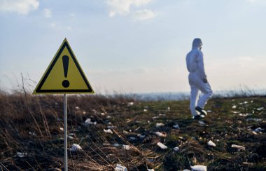 Tehlike ve tehlikelerle ilgili sarı üçgen uyarı işareti. Bulanık arka planda radyasyon kıyafeti giyen çevreci. Ekoloji, çevre kirliliği ve tehlike kavramı