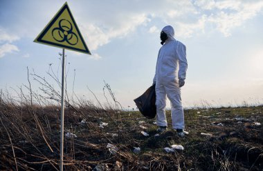 Koruyucu giysili ve gaz maskeli bir çevreci çöp torbası tutuyor, çöplerle dolu terk edilmiş bölgede biyolojik tehlike işaretinin yanında duruyor, başka tarafa bakıyor. Ekoloji ve biyolojik tehlike kavramı.