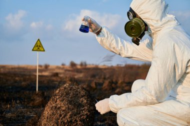 Takım elbiseli bir ekolojist, gaz maskesi elinde mavi sıvıyla test tüpü tutarken yanmış topraklarda biyolojik tehlike işareti olan yanmış ot ve toprağı inceliyor. Çevreci, yangın üstüne laboratuvar testi yapıyor.