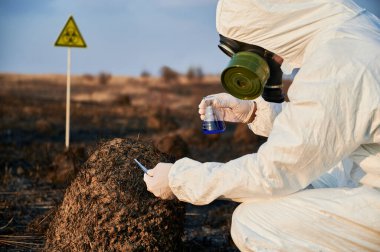 Çevreci, yangın üstüne laboratuvar testleri yapıyor koruyucu giysi, gaz maskesi takıyor, yanmış topraklarda biyolojik tehlike işareti ile yanmış çimenleri ve toprağı inceliyor. Ekoloji ve araştırma kavramı