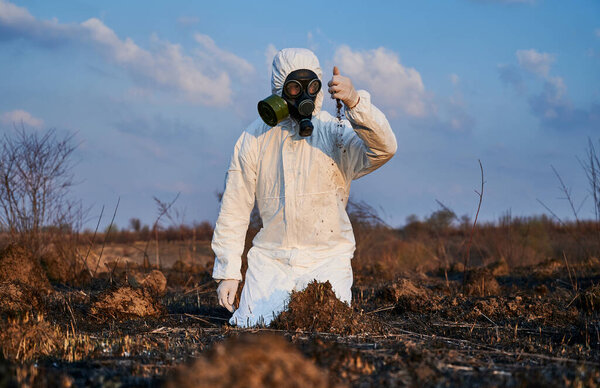 Male environmentalist in suit and gas mask pouring out soil from his hand while doing research work on scorched territory, analyzing condition of soil. Concept of ecology, research and burned earth.