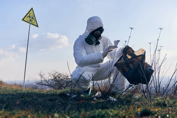 Ecologist putting piece of dirty used paper into garbage bag. Male research scientist in protective suit, gas mask picking up garbage in field with biohazard sign. Concept of environmental pollution