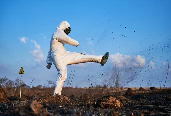 Disappointed scientist, wearing a white protective suit, gas mask and gloves, kicking soil on scorched field against blue sky. Concept of ecology and burned earth.