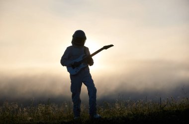 Arka planda beyaz mistik gökyüzü olan sisli bir vadide gitar çalan bir uzay gezgini silueti. Uzay giysisi ve miğfer takarken astronot geldi. Müzik, astronotluk ve doğa kavramı.