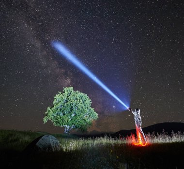 Uzay yolcusu görkemli gece yıldızlı gökyüzünü fenerle aydınlatıyor. Beyaz uzay giysili kozmonot, çadır ve ağaçla otlak vadide dikiliyor. Galaksi kavramı, uzayda insan ve Samanyolu.
