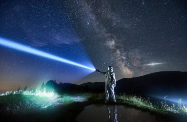 Astronot fenerle muhteşem gece gökyüzünü aydınlatıyor. Kozmonot, çimenli vadide küçük bir gölün yanında dururken beyaz uzay giysisi ve kask giyiyor. Işık, galaksi ve astronotlar kavramı.