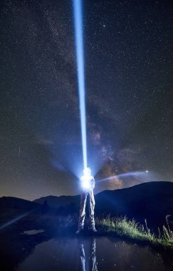 Uzay giysisi giyen kozmonot, göl kenarındaki yıldızlı gökyüzüne mavi ışık hüzmesi yolluyor, bacakları suya yansıyor. Işık, galaksi ve astronotlar kavramı.