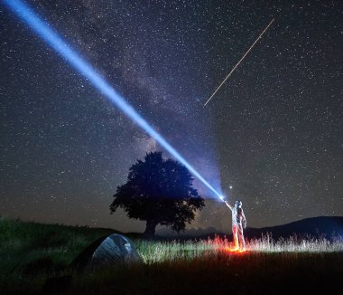Uzay giysisi ve miğferiyle aydınlanmış uzay adamı turist kampı yakınlarındaki fenerlerin yardımıyla yıldızlarla dolu gece gökyüzüne bir ışık hüzmesi yolluyor. Turist çadırı, büyük ağaç, arka planda kayan yıldız.