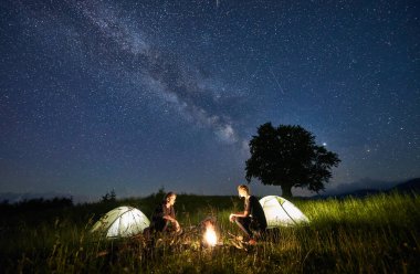 Güzel bir gece manzarası çayırların üzerindeki yıldızlı gökyüzü aydınlanmış kamp çadırlarının yanındaki yürüyüşçülerle. Turistler, yıldızlı mavi gökyüzünün altında kamp ateşinin yanında oturuyorlar. Seyahat, yürüyüş ve kamp kavramı.