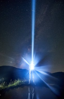 Uzay giysisi giyen astronot, göl kenarındaki yıldızlı gökyüzüne mavi ışık hüzmesi yolluyor, bacakları suya yansıyor. Uzay yolculuğu kavramı