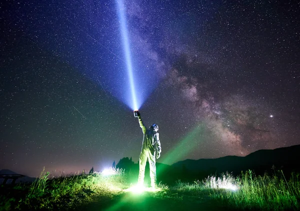 Spaceman wearing white space suit and helmet directs a blue ray of light into sky full of stars with a help of lantern in the night in the mountains, standing on illuminated green grass. Space concept
