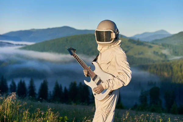 宇航员身穿白色宇航服 头戴白色吉它的头盔 站在阳光灿烂的绿色雪地上 背景是雾蒙蒙的晨山奇景 — 图库照片