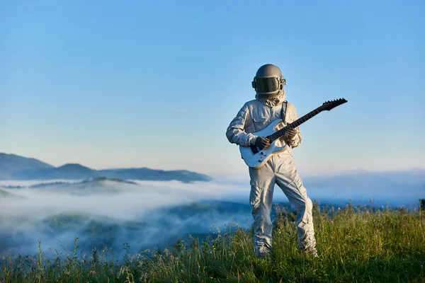 Astronaut wearing white space suit and helmet playing white guitar, standing on sunny green mountain glade in summer under blue sky, morning fog rising up from the valley behind him.