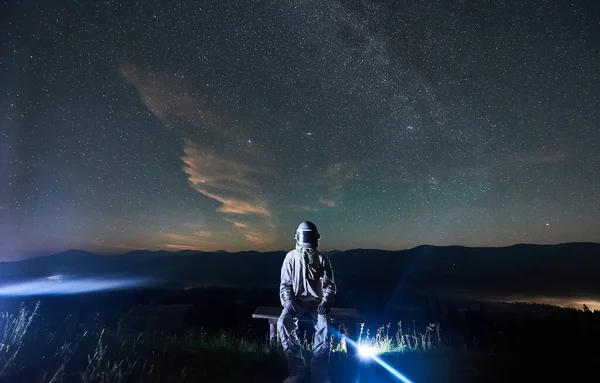 Illuminated astronaut wearing white space suit and helmet sitting on bench on hill at night. Mountain ridge and sky full of stars on background. Concept of space travel, galaxy and cosmonautics.