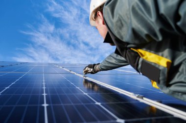 Güvenlik kaskı, üniforma ve çalışma eldiveni takan elektrikçi, arka planda mavi gökyüzü olan uğursuz anahtarın yardımıyla parlak yeni bir güneş pili yerleştiriyor. Alternatif yeşil enerji kaynakları ve yenilikler kavramı.